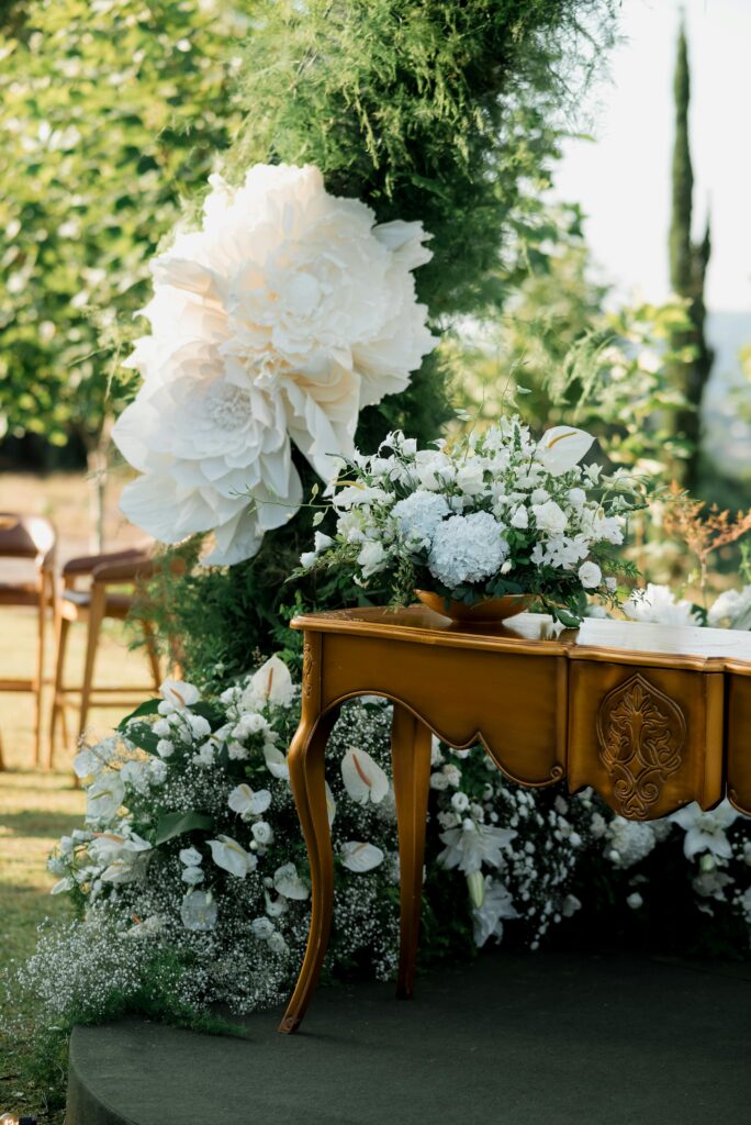 A wooden table decorated with white floral arrangements and draping in front of green foliage.