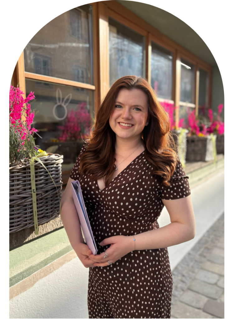 Georgia Tamlyn wearing a brown polka dot dress stands outside holding a folder, with planters and pink flowers in the background.