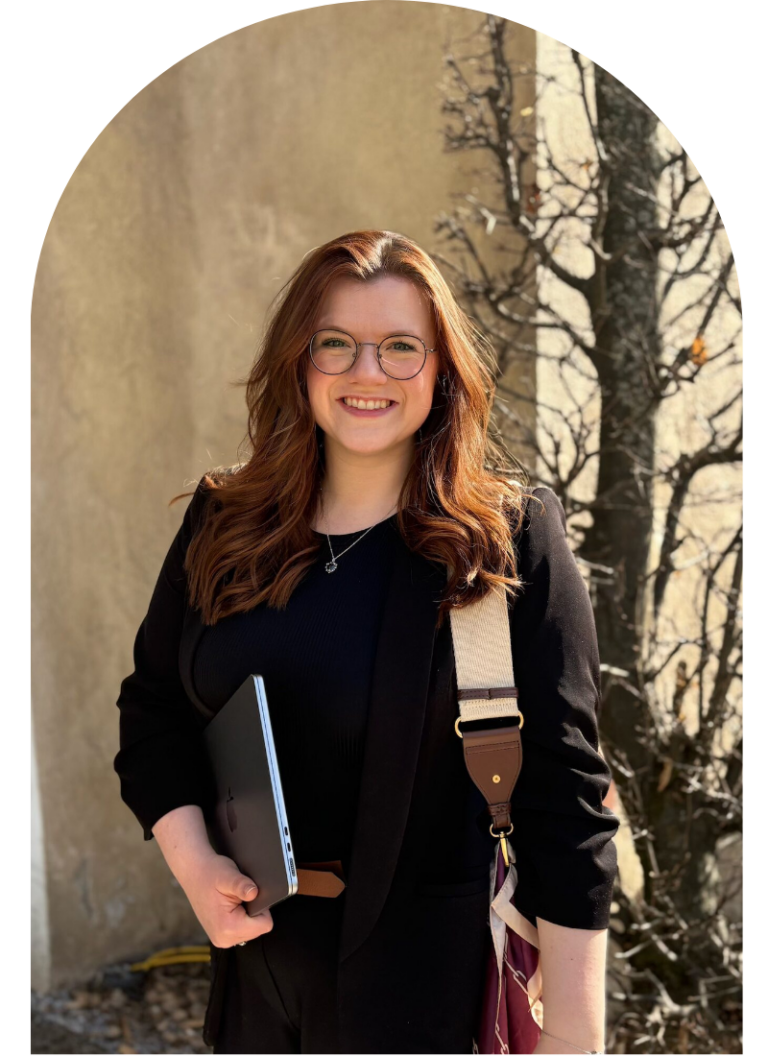 Georgia Tamlyn, smiling and standing outdoors in sunlight against a pale wall, holding a laptop or notebook, with a shoulder bag strap visible and tree shadows cast behind her.