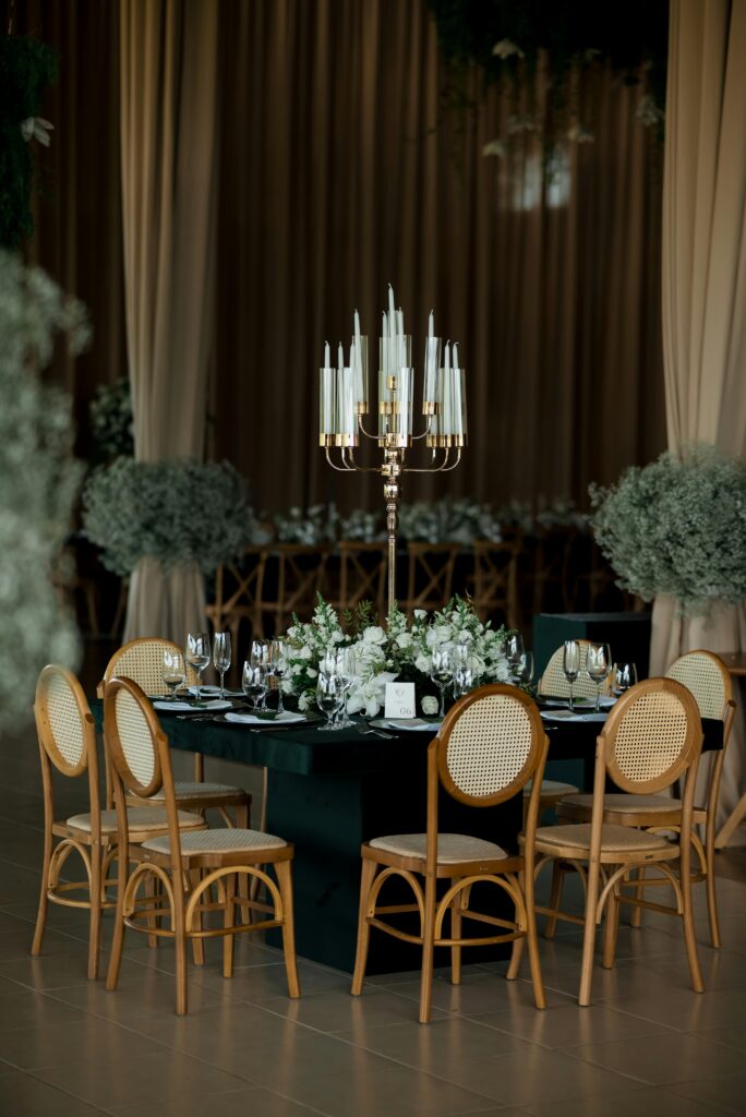 A formal dining table with dark green cloth, brass candelabra with white candles, white floral centrepiece, wooden rattan chairs, and grey-green foliage arrangements, set against draped curtains.