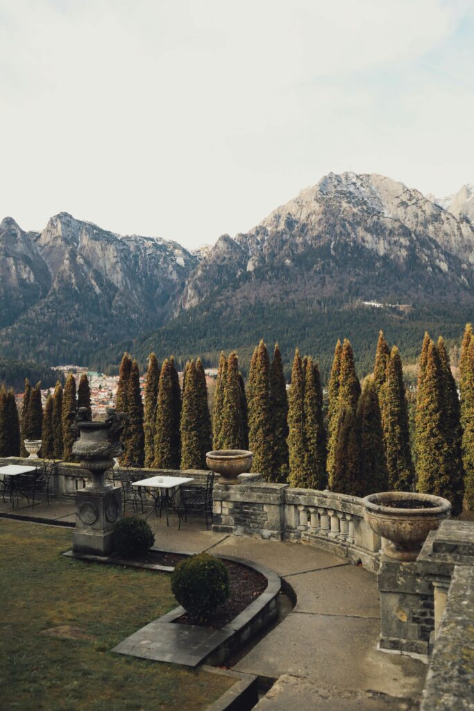 A terrace with cypress trees and stone urns overlooks a valley with snow-capped mountains and a distant village nestled among forested peaks.